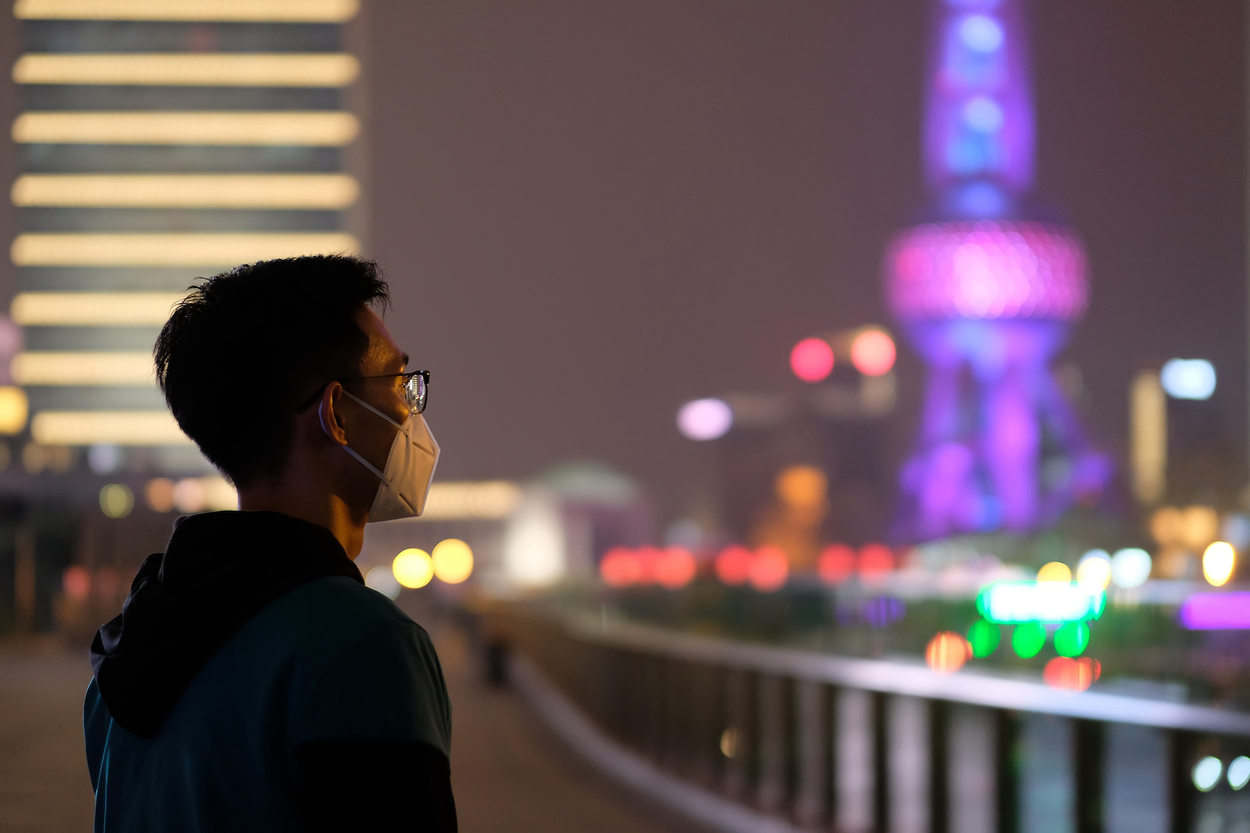 A young person in Shanghai stands alone wearing a medical face mask and looks off at the colourful skyline