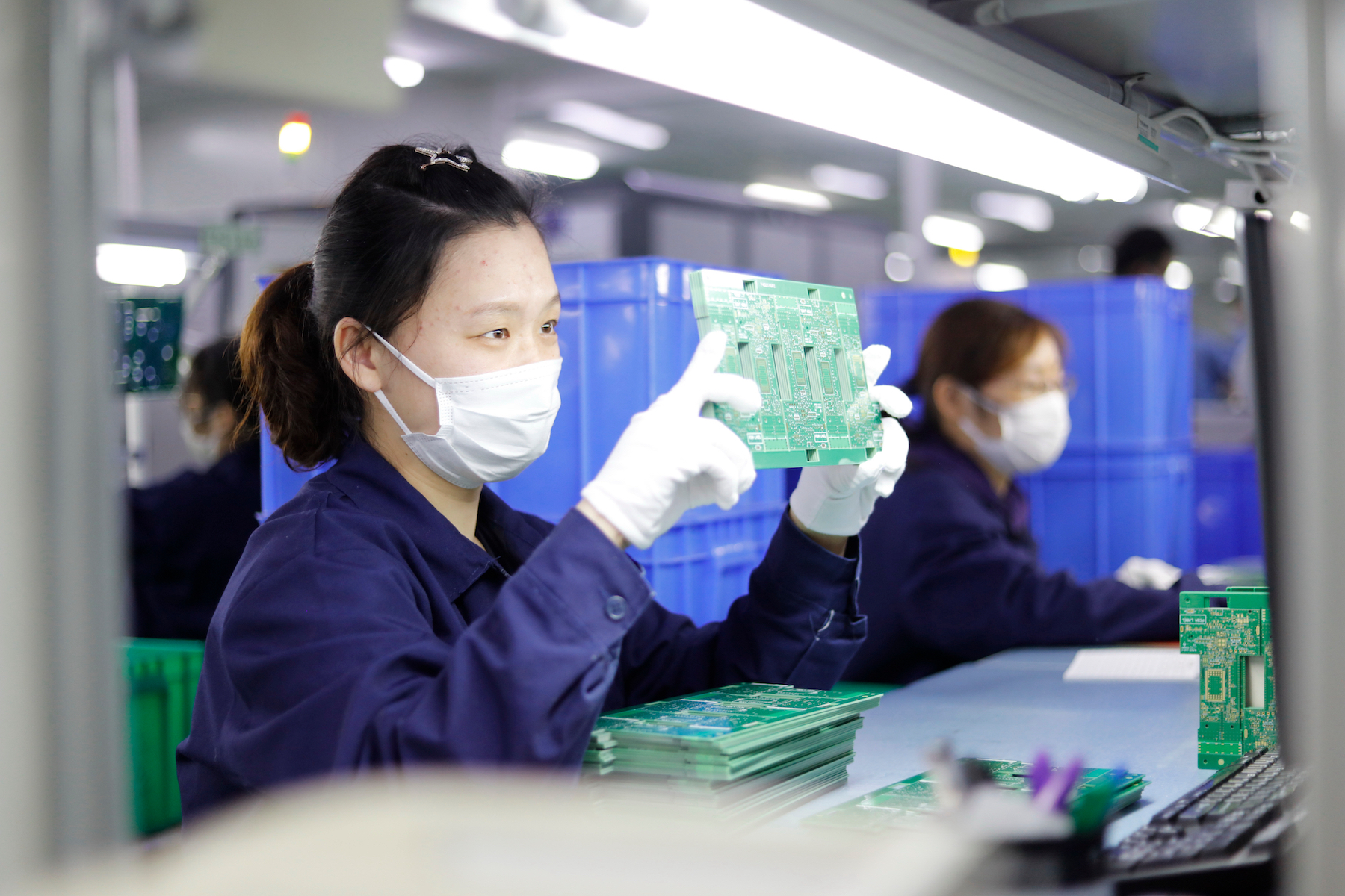 A female factory worker wearing a medical mask inspects a piece of electronics