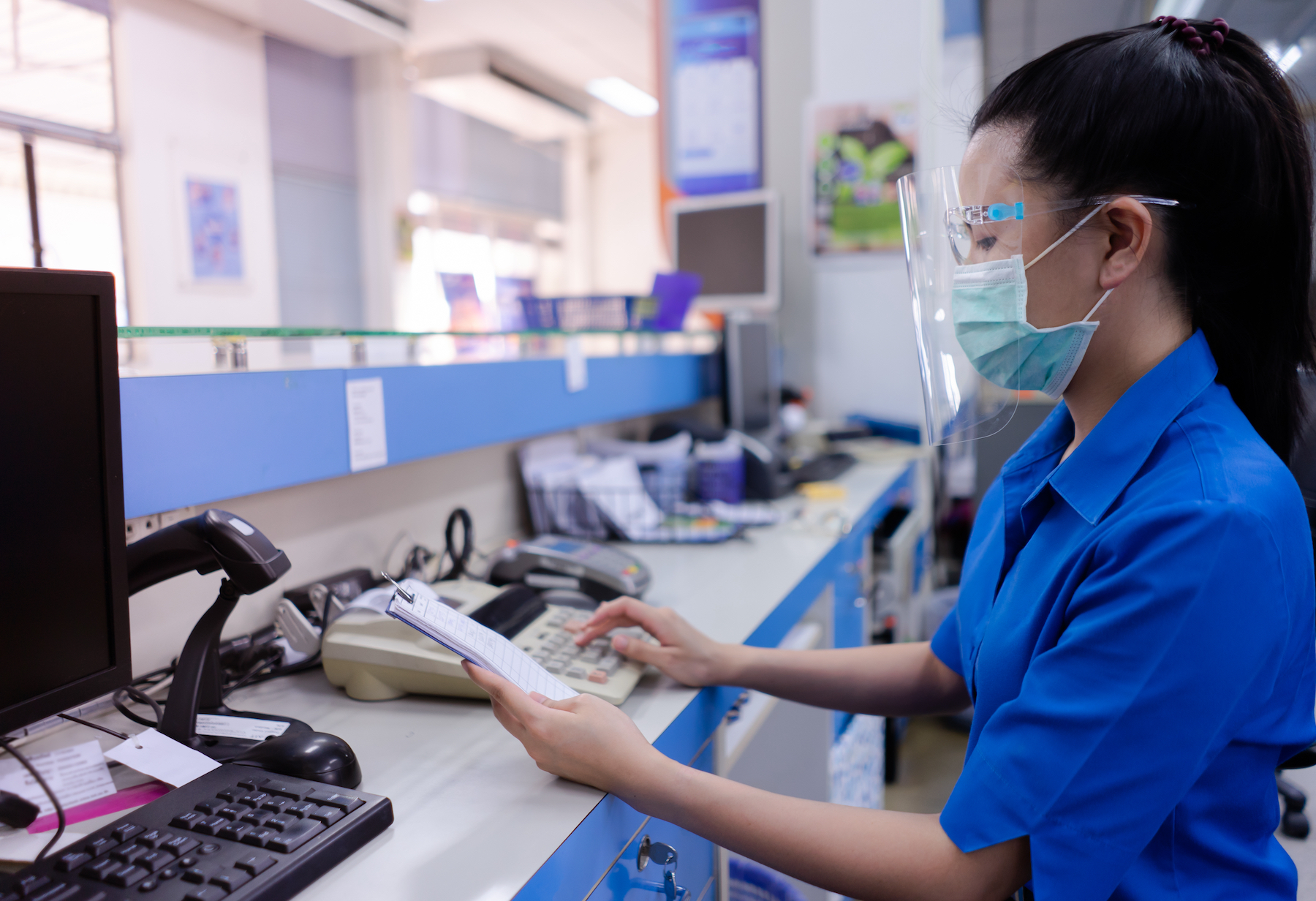 A woman office worker sits at a computer and wears a clear face shield