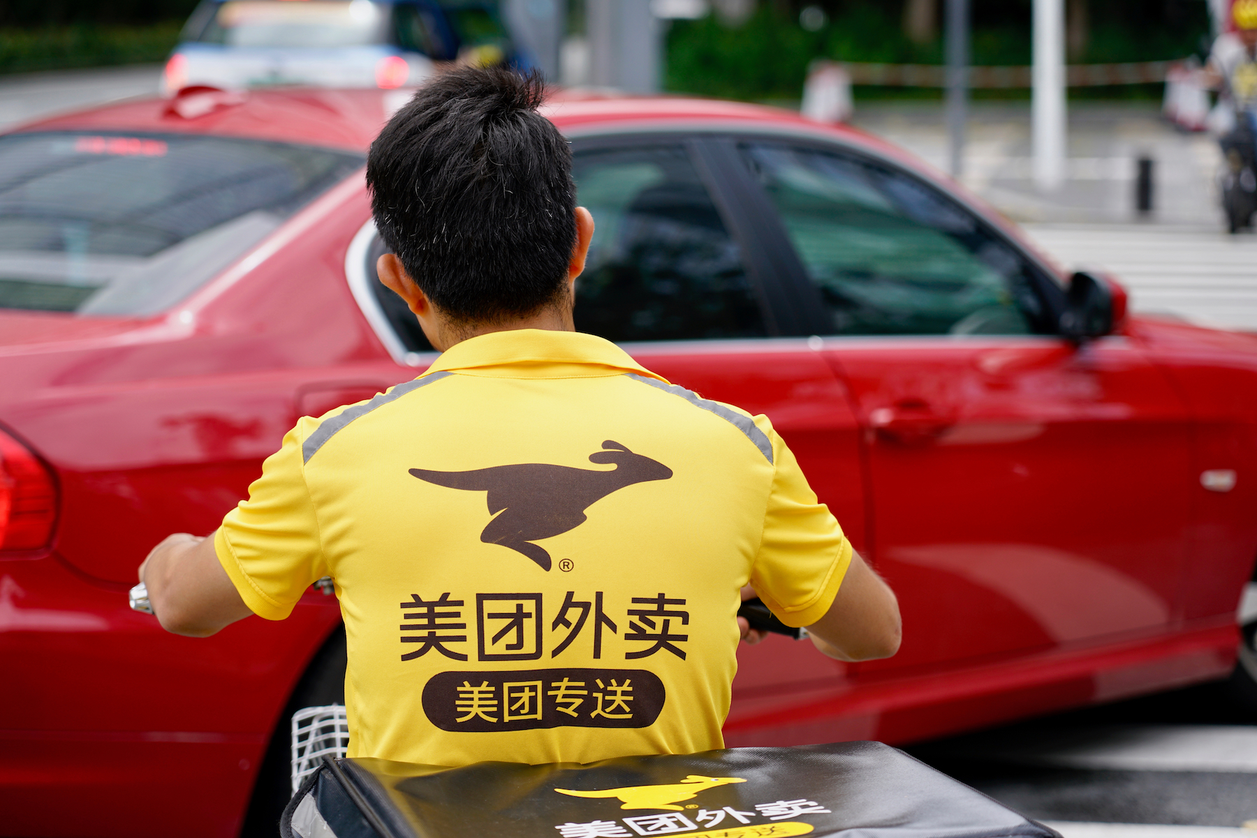 A Meituan food delivery rider drives in traffic behind a red car