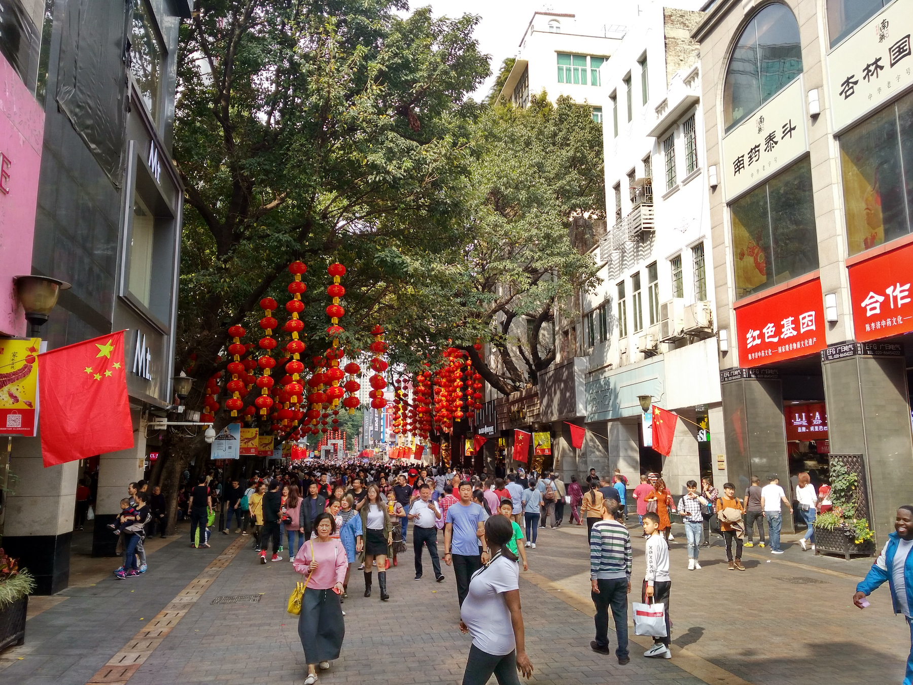 A pregnant woman in the foreground of the photo walks across a busy pedestrian shopping street