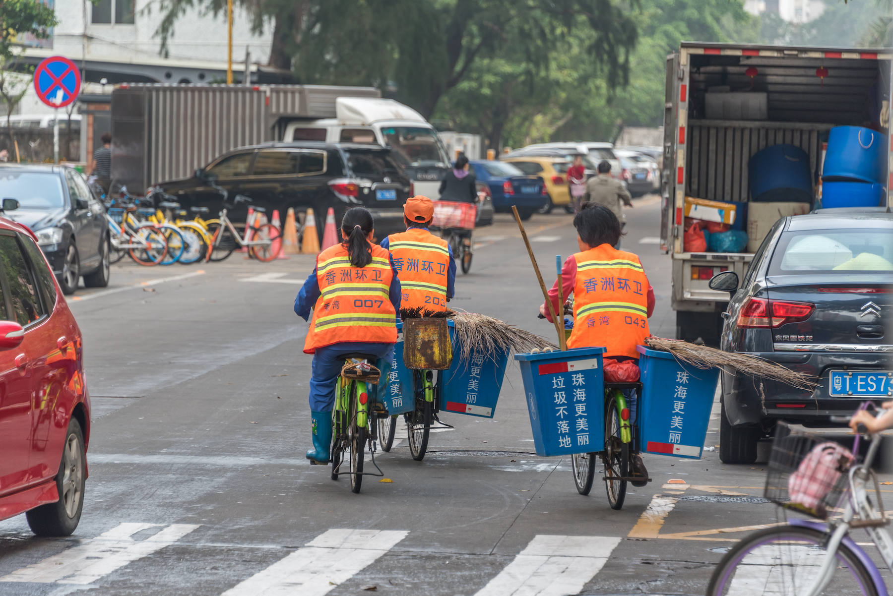 Three sanitation workers on bicycles with blue plastic bins attached to either side ride in the city streets wearing orange vests and carrying brooms