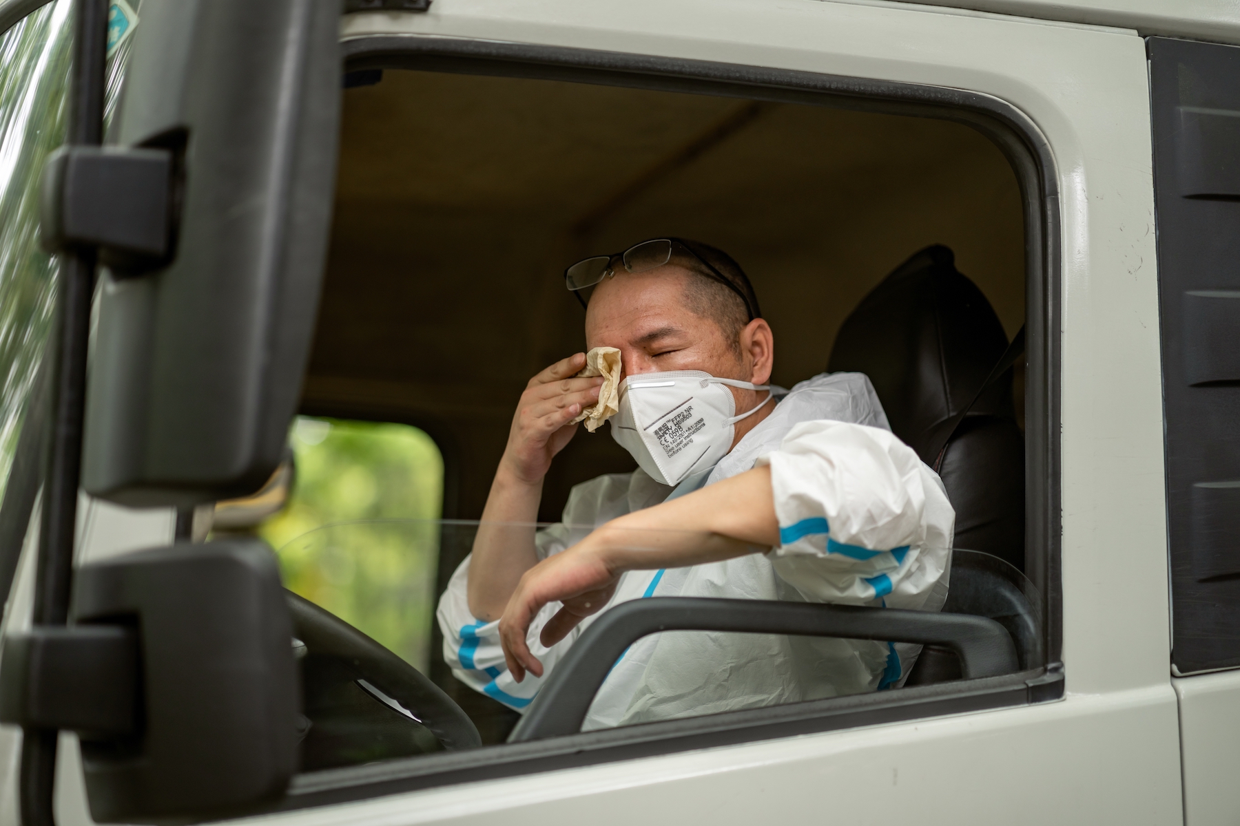 A truck driver wearing a face mask and in full PPE wipes his face while seated in the cab of a truck